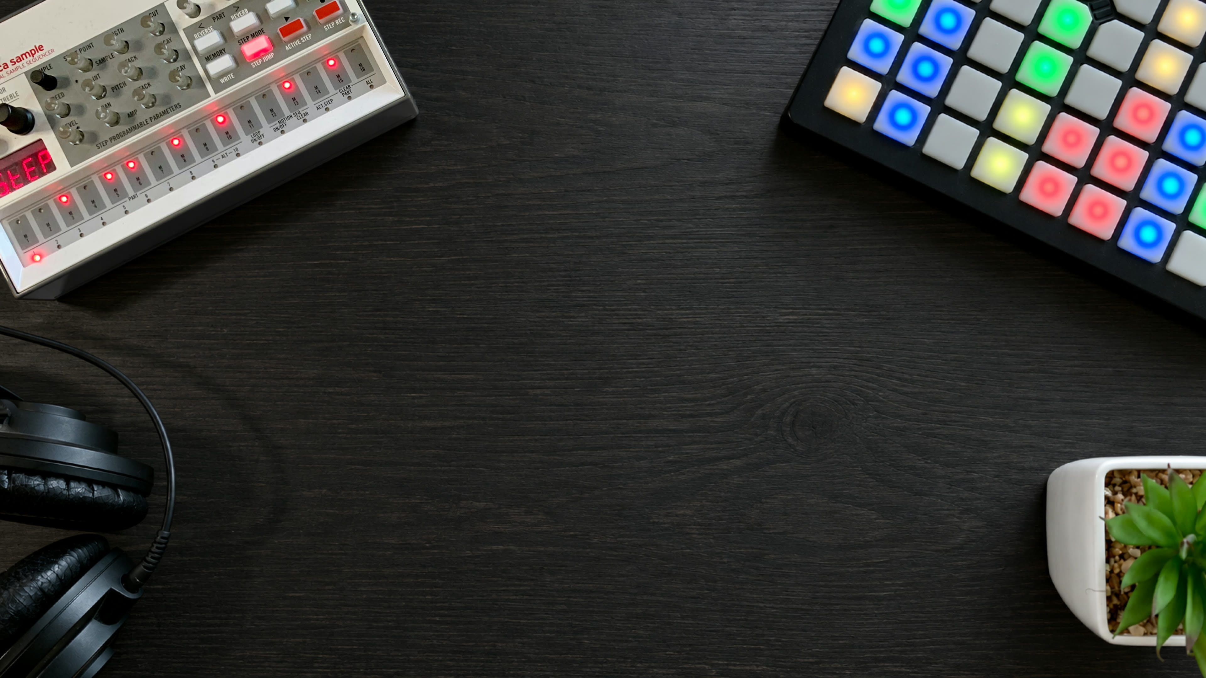 A music studio desk: a sampler, a lit pad controller, headphones and a small plant around dark wood